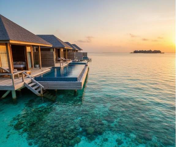 두짓 타니 몰디브의 워터빌라들이 석양을 배경으로 에메랄드빛 바다 위에 늘어서 있으며, 개인 인피니티 풀 아래로 선명한 산호초와 열대어가 보인다.