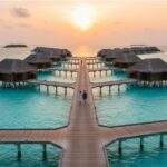 몰디브 신혼여행: 리조트의 워터빌라들이 석양 아래 푸른 바다 위에 길게 늘어서 있고, 다리 위로 한 커플이 걸어가는 모습.