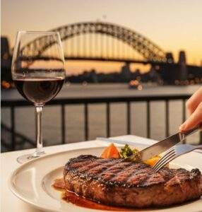 시드니 하버 브리지를 배경으로 야외 레스토랑에서 맛있는 스테이크와 와인을 즐기는 사람의 시점 이미지