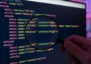 컴퓨터 모니터 화면의 HTML 코드를 돋보기로 확대하여 <meta name="revenue" content="high"> 태그를 검사하는 손 클로즈업.