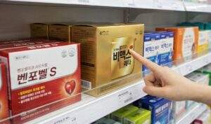 성남 메가팩토리 약국 진열대에 비맥스 메타와 벤포벨 S 영양제 박스가 나란히 놓여 있고, 소비자가 비맥스 메타의 성분표를 손가락으로 가리키며 비교하고 있다.